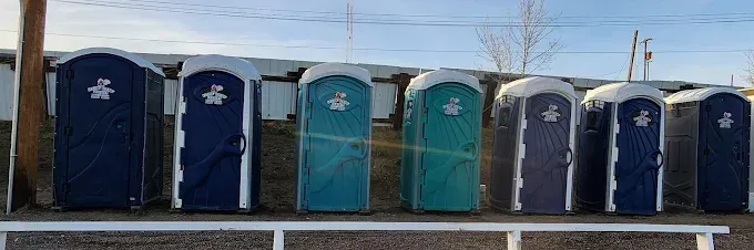 A row of seven portable toilets in shades of blue and teal, positioned side-by-side on a dirt lot outdoors.