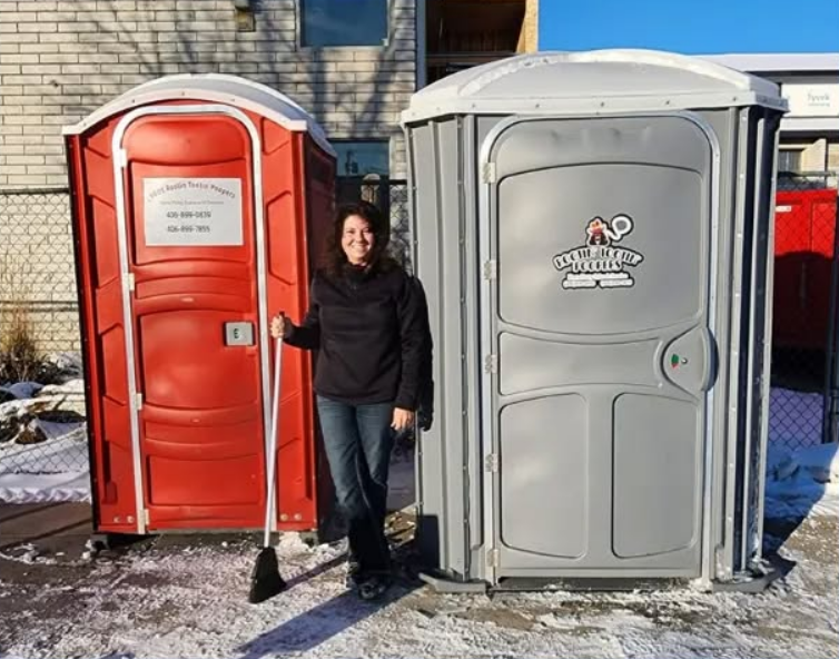 A person stands between a red portable toilet and a grey portable toilet outside on a snowy day, holding a broom.