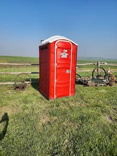 A red portable toilet stands on a grassy field in front of a wooden fence and rustic wagon wheels under a clear blue sky.