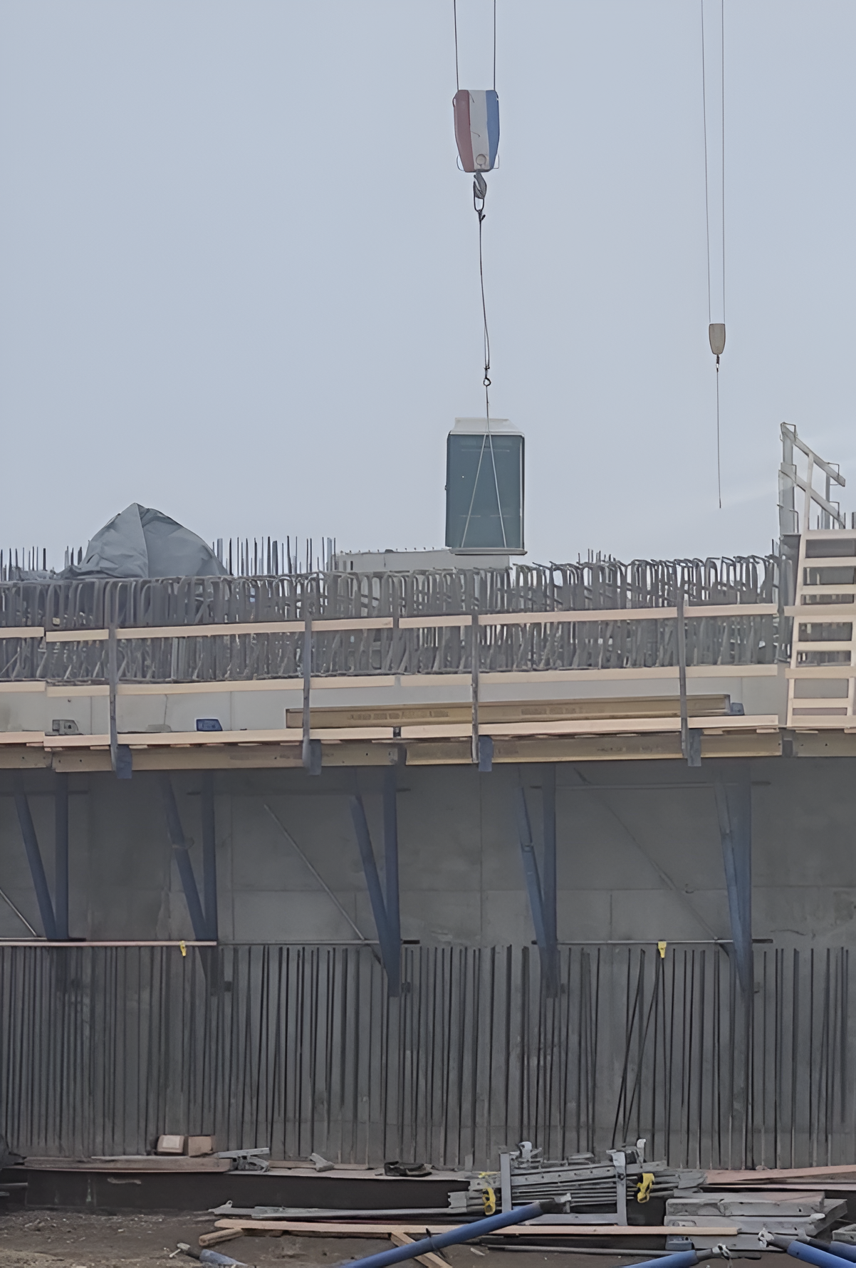 A crane lowers a rectangular green component onto a concrete construction site with exposed rebar and scaffolding.