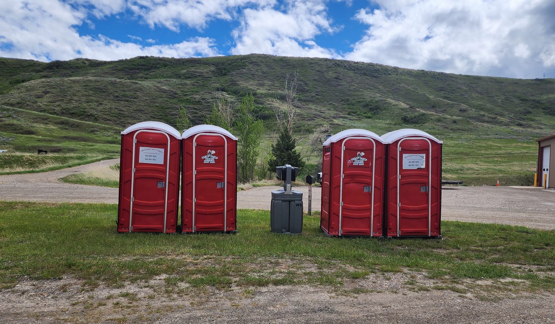 Four red portable toilets stand in two pairs on a grassy field in front of a hillside.