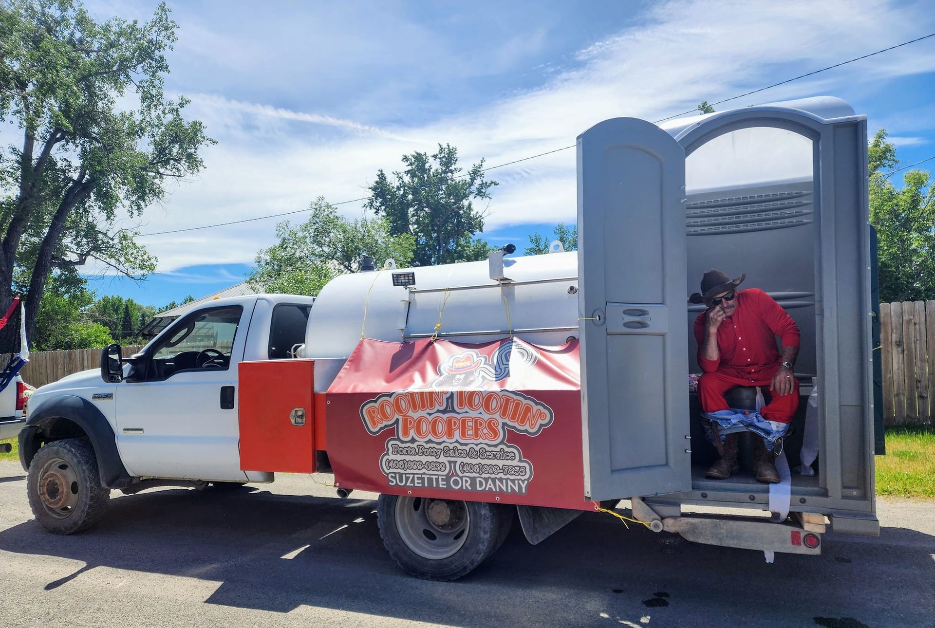 A white truck features a red banner and a portable toilet on the bed, where a person in a red outfit is seated inside.