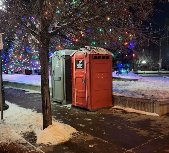 Two portable toilets, one gray and one red, sit on a snow-covered sidewalk beneath a tree decorated with holiday lights.