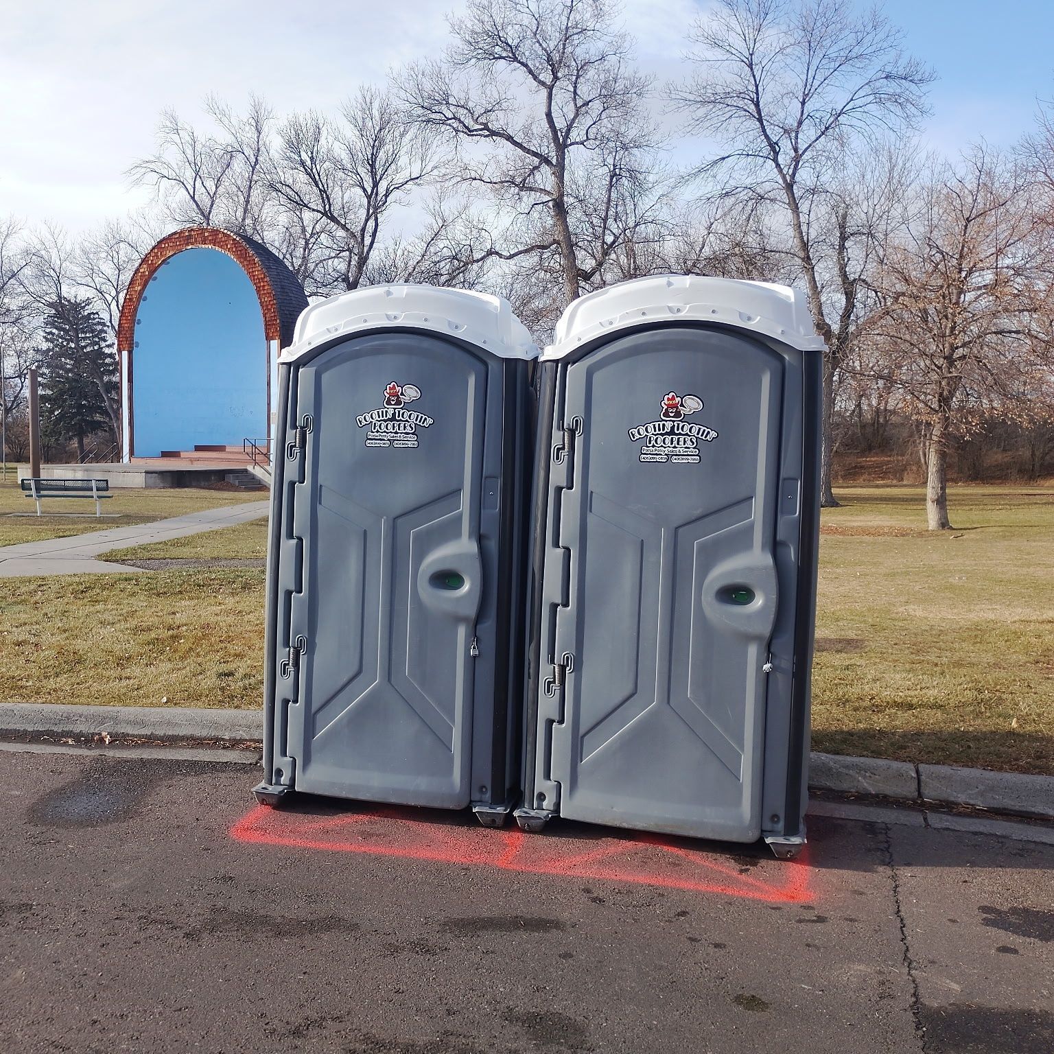 Two grey portable toilets sit on a paved area with red markings, positioned in a park near a blue-arched structure.