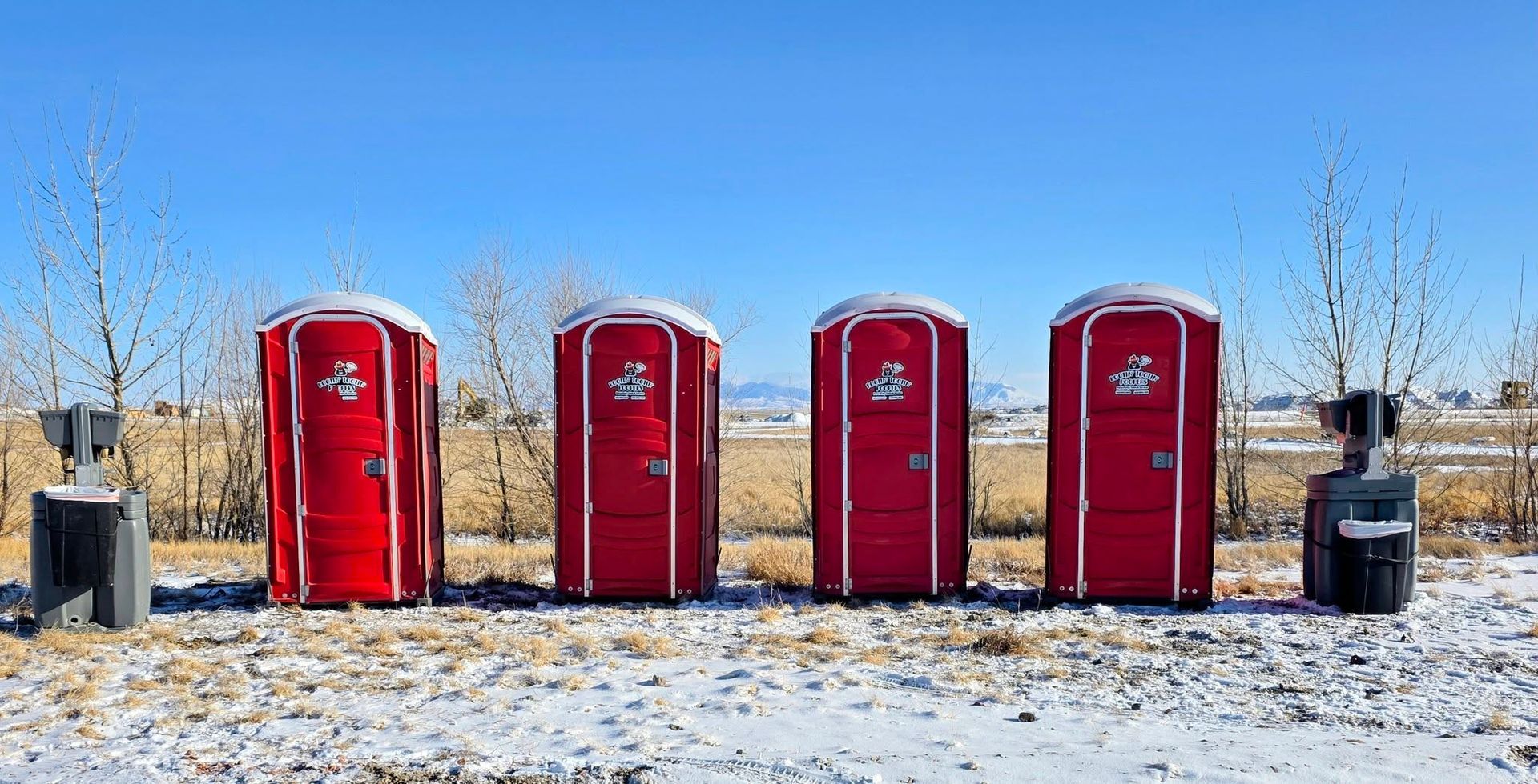 Four red portable toilets in a row outdoors on a snowy, sunny day with sanitation stations on either side.