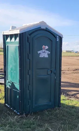 A dark green portable toilet stands on a dirt field under a clear blue sky.
