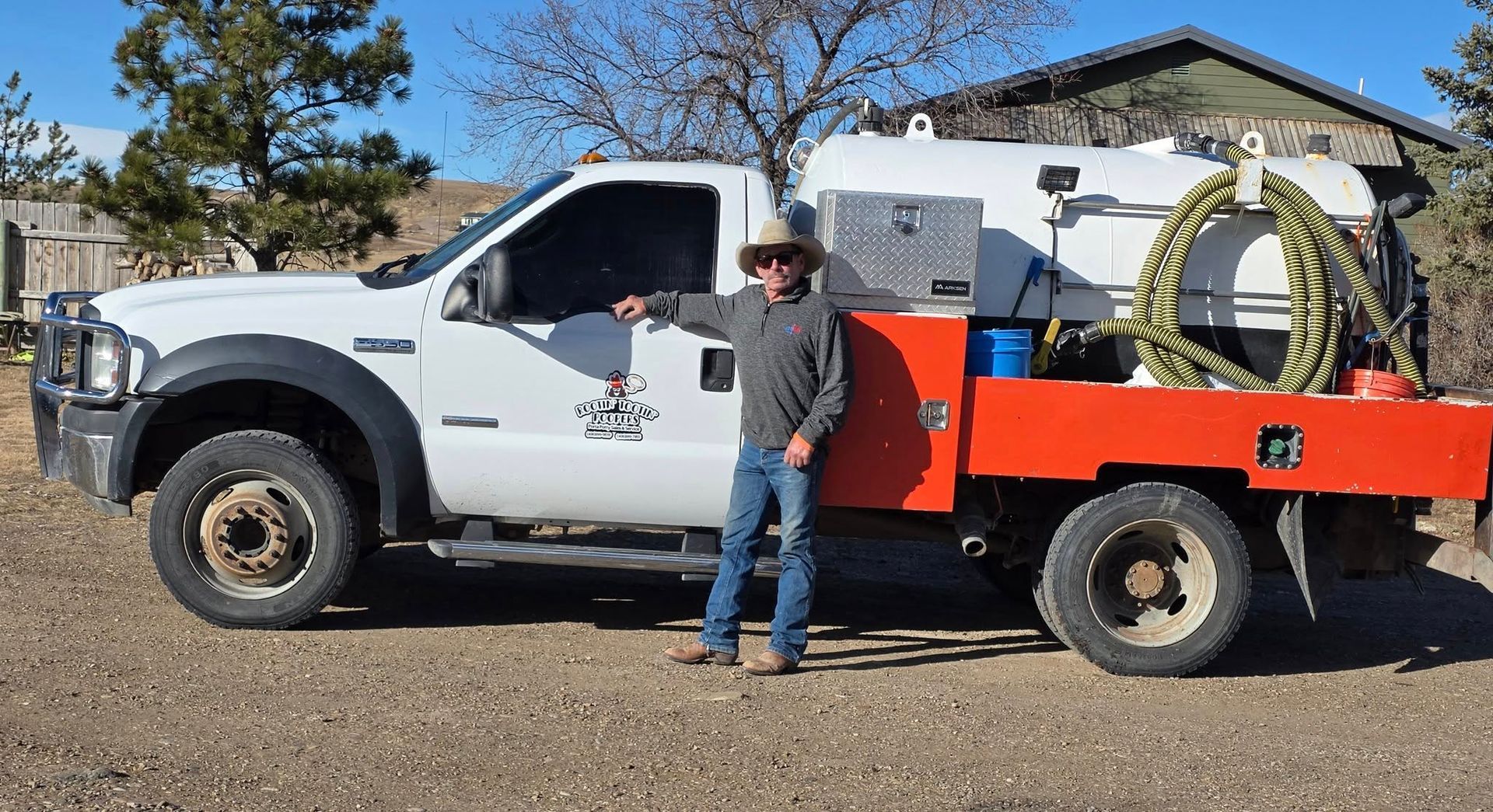 A person in a cowboy hat stands by a white work truck with an orange flatbed and a large cylindrical tank, parked outdoors.