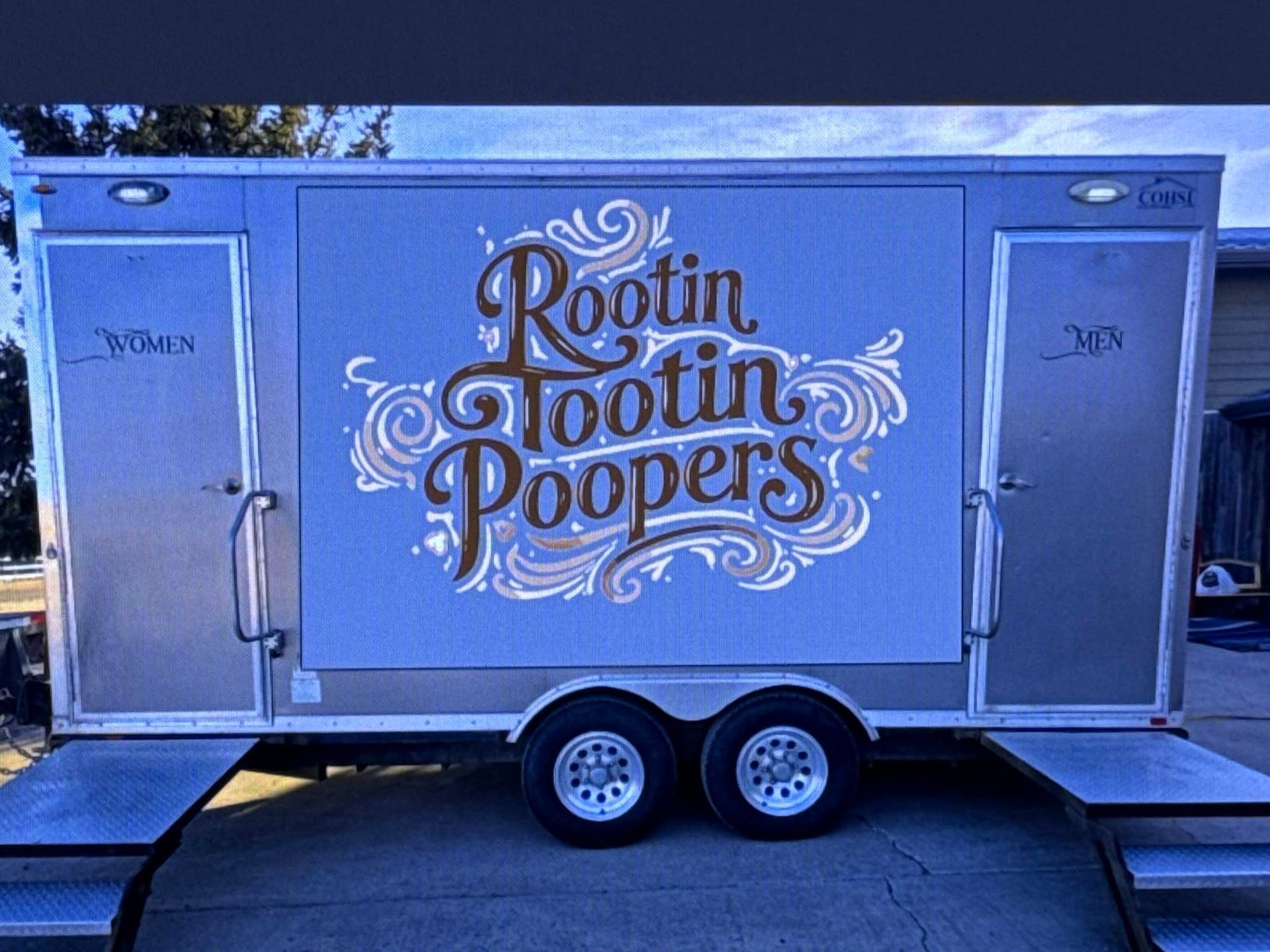 A portable restroom trailer with silver walls, two doors, and a stylized brown logo reading
