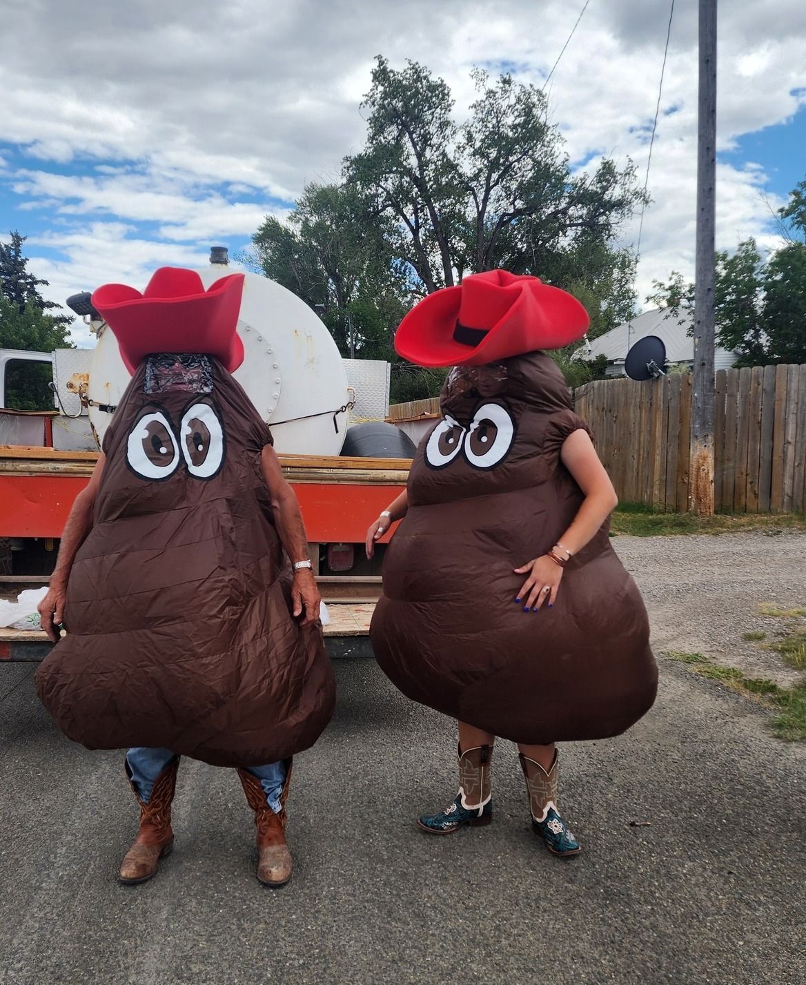 Two people wearing brown poop-shaped costumes and red cowboy hats standing outdoors.