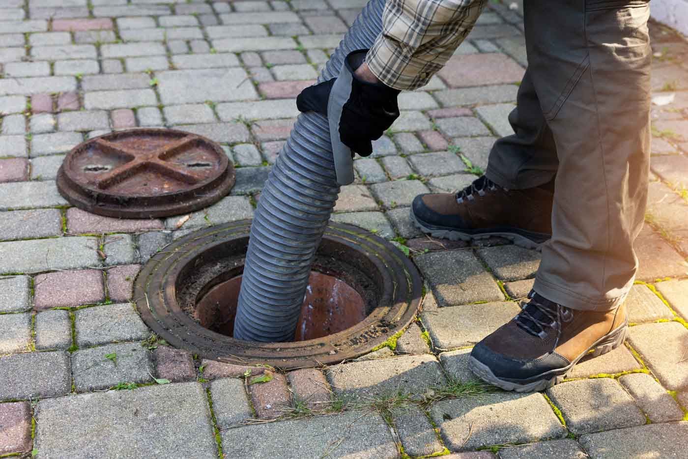 A person in work clothes uses a large suction hose to clean out an open residential sewer access port on a paved patio.