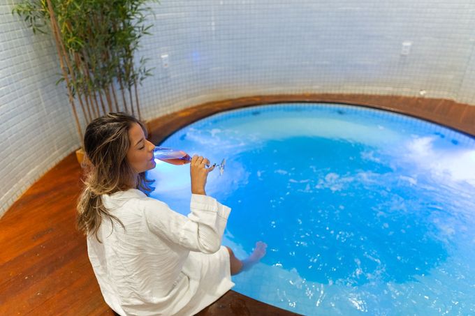 Uma pessoa vestida com um robe branco está sentada à beira de uma piscina circular iluminada em azul, bebendo de um copo com os pés mergulhados na água.