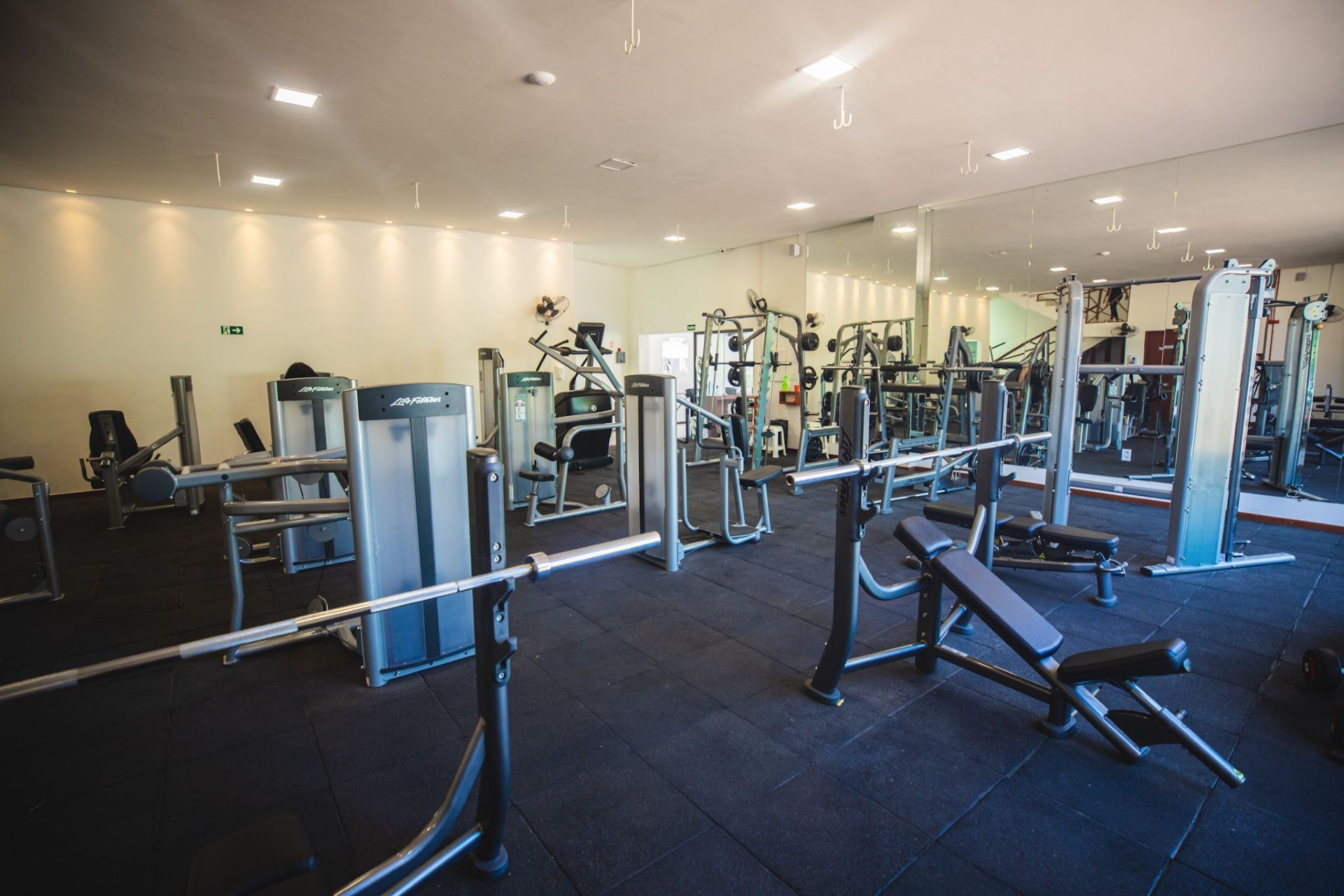 Academia vazia com bancos de musculação, racks e diversos aparelhos de musculação sobre piso de borracha preta.