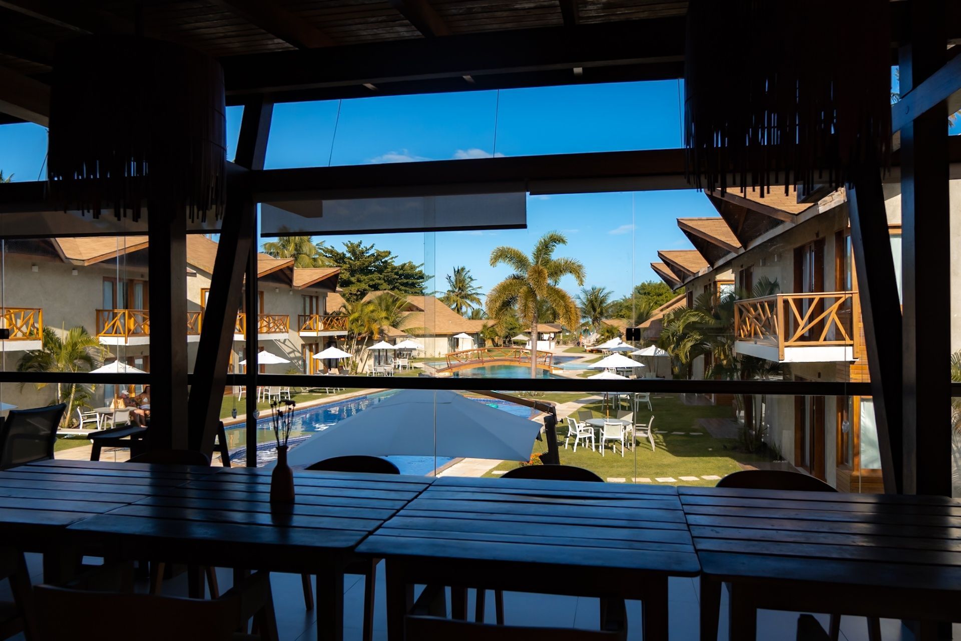 Vista de uma área de jantar interna sombreada com vista para um pátio ensolarado do resort, com piscina e vilas do hotel.
