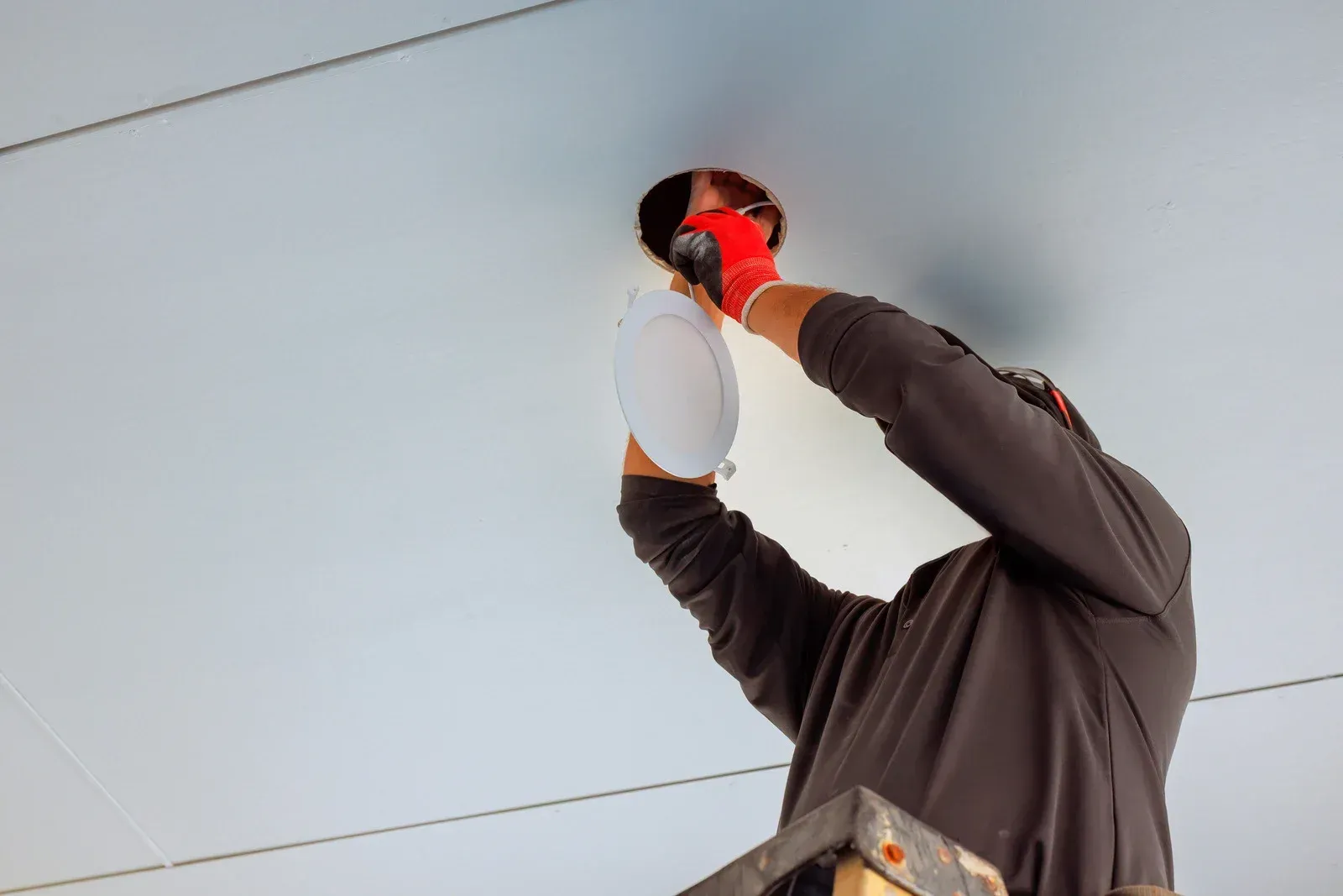 Worker installing a ceiling light while standing on a ladder, holding a white fixture overhead