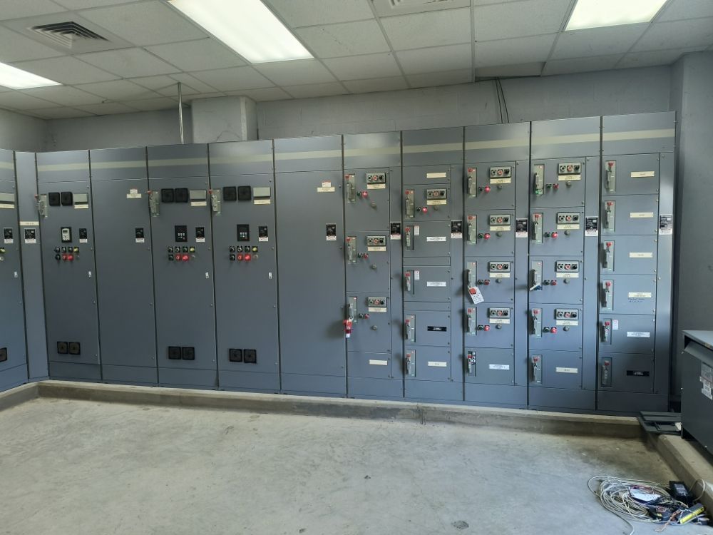 Row of gray electrical control panels in an industrial room with a concrete floor and fluorescent lights.