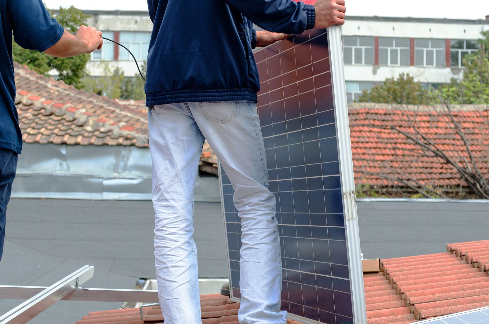 Two workers installing a solar panel on a rooftop, with red-tiled roofs in the background