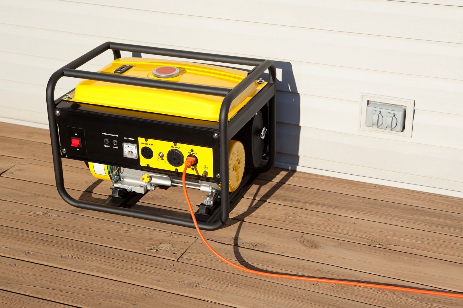 Yellow portable generator in a black frame on a wooden floor, with cords plugged in against a wall.