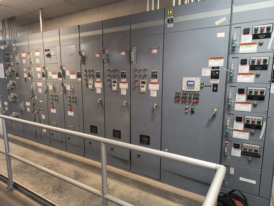 Industrial electrical control panels in a facility, with gray metal cabinets and gauges behind a railing.