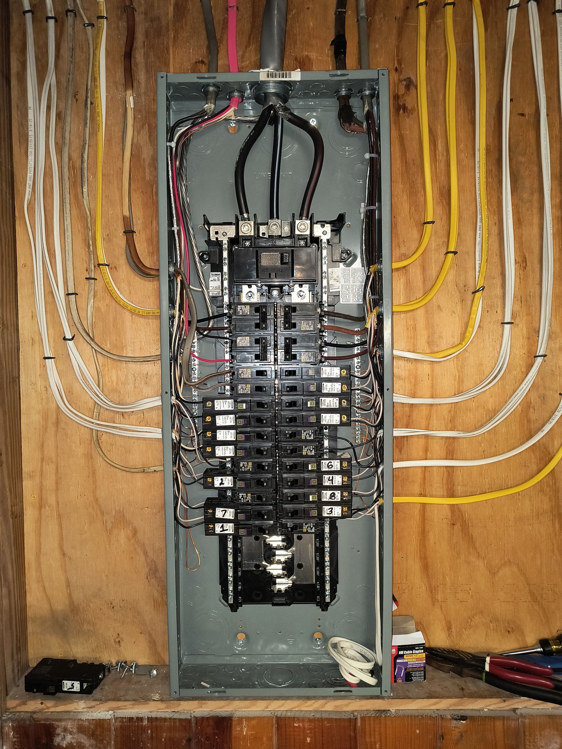 Open electrical breaker panel with many black switches and colored wires inside a wooden wall cavity