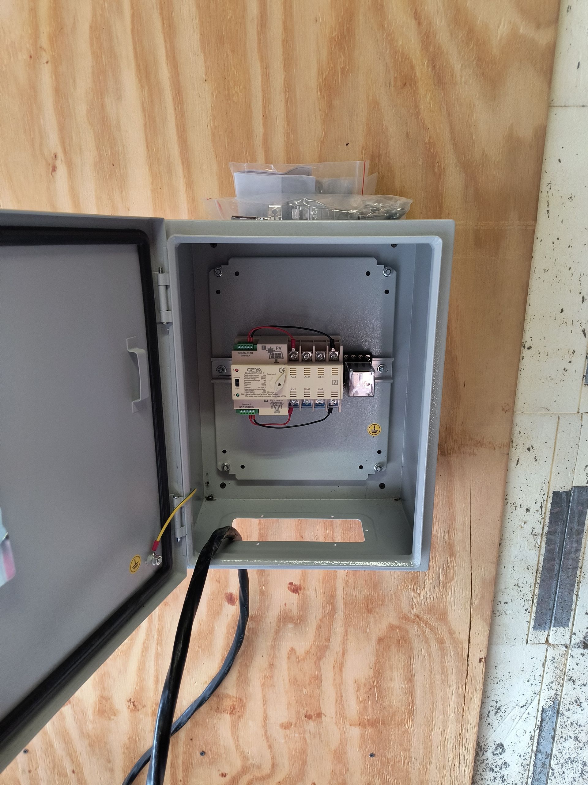 Open electrical breaker panel mounted on plywood wall with visible wiring and label tags