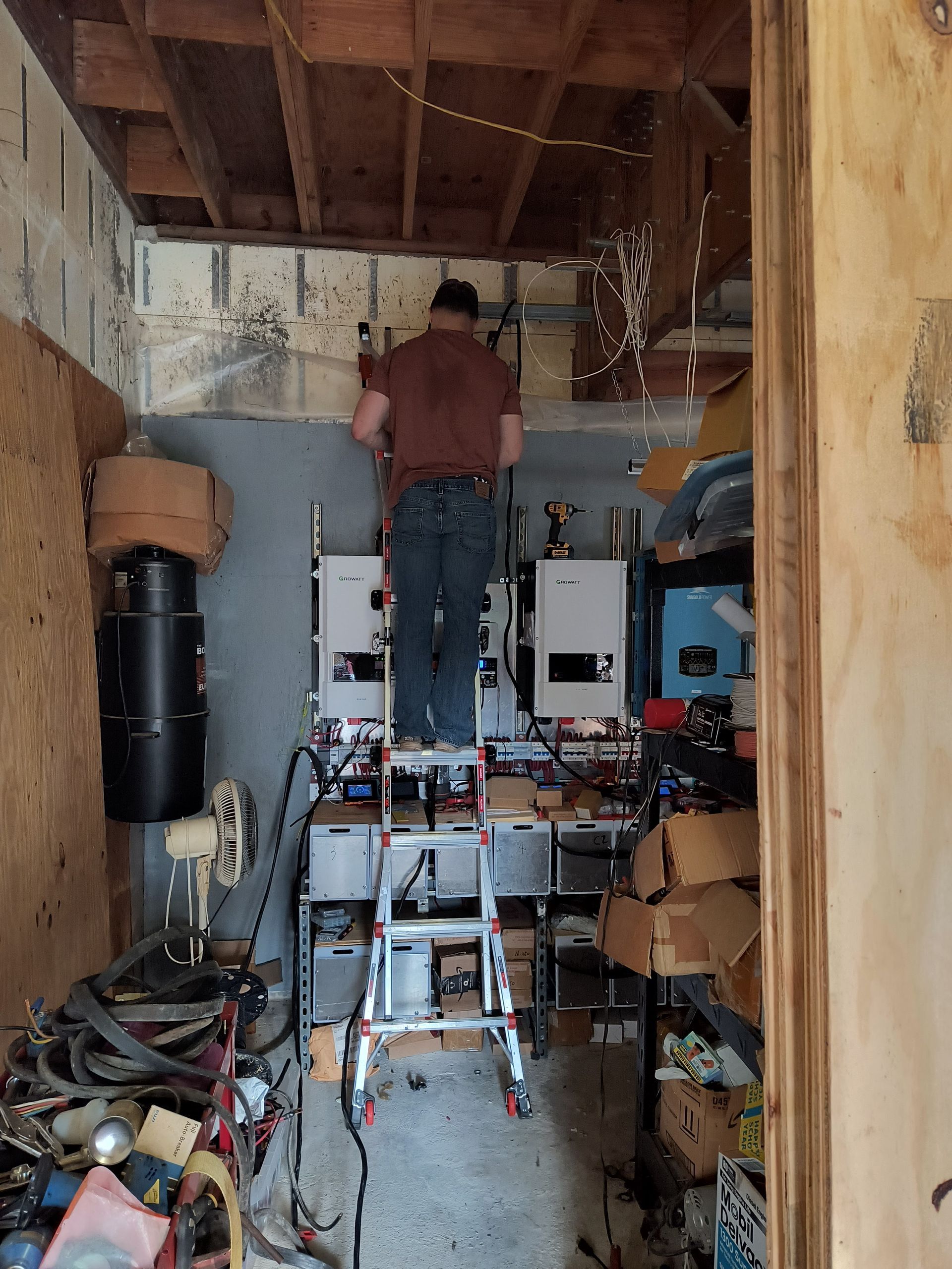 Worker on a ladder repairing wiring in a cluttered attic with exposed beams and tools.