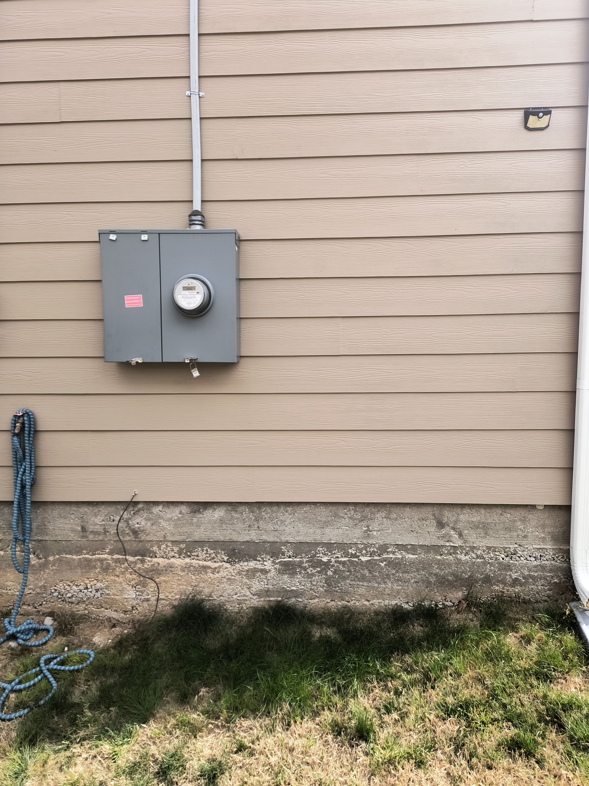 Gray electrical meter box on beige house siding above a gravel foundation and grass strip.