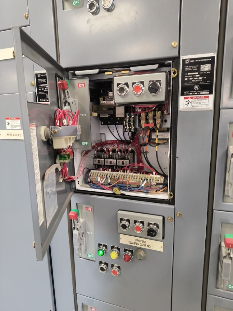 Open electrical control cabinet with wiring, relays, and illuminated push buttons in an industrial setting