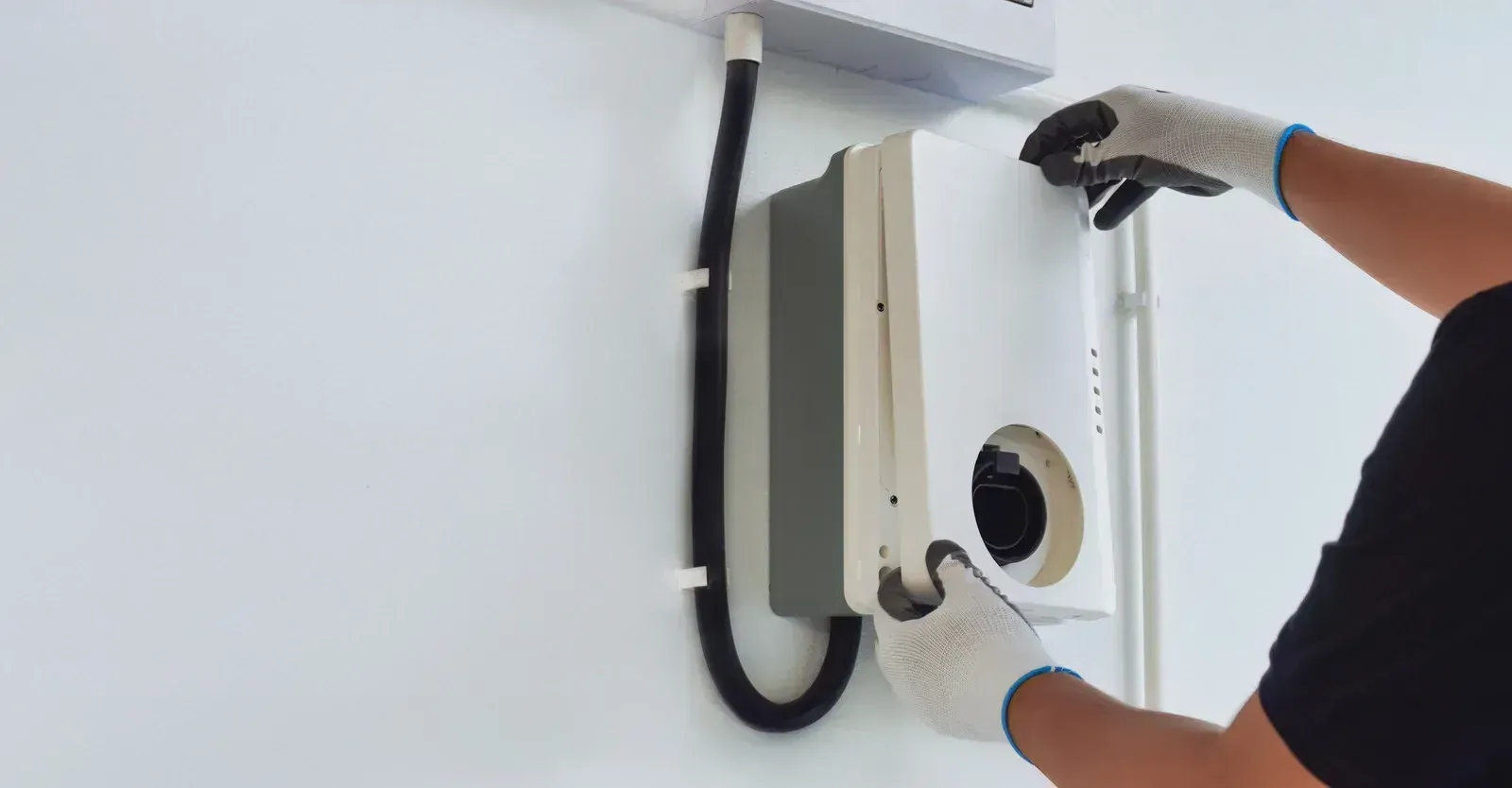 Gloved hands adjust a white wall-mounted device with a circular opening on a light gray wall.