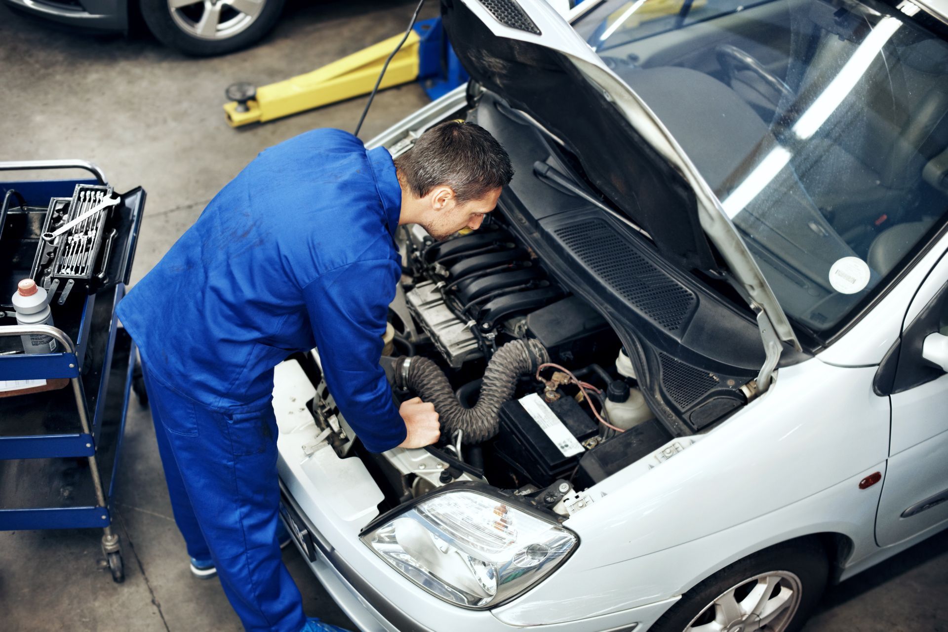 Mechanic in blue coveralls inspecting a white car’s engine with the hood open