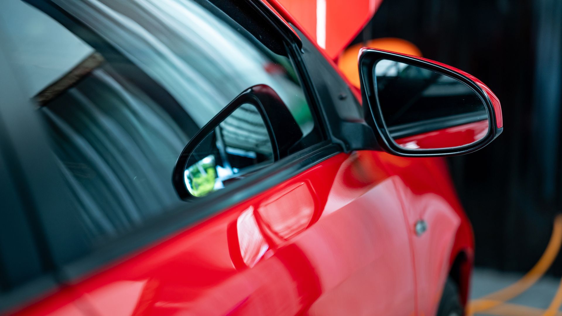 Close-up of a red car’s side mirror and door with reflections in a blurred garage setting