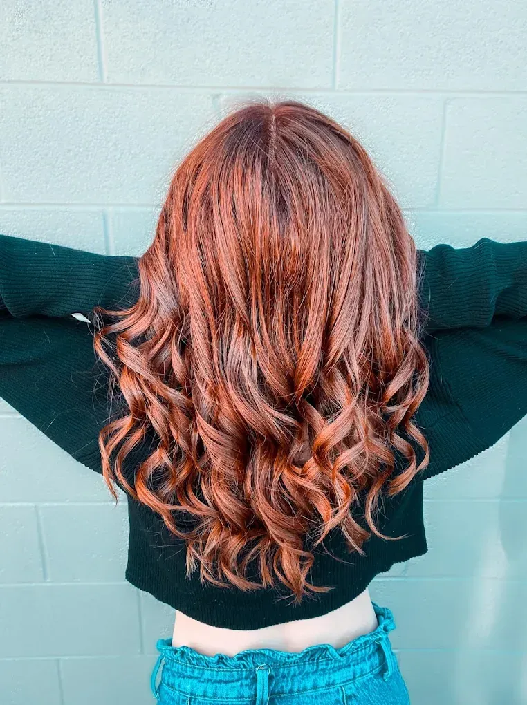 Person with long wavy auburn hair seen from behind, wearing a black top and blue jeans.