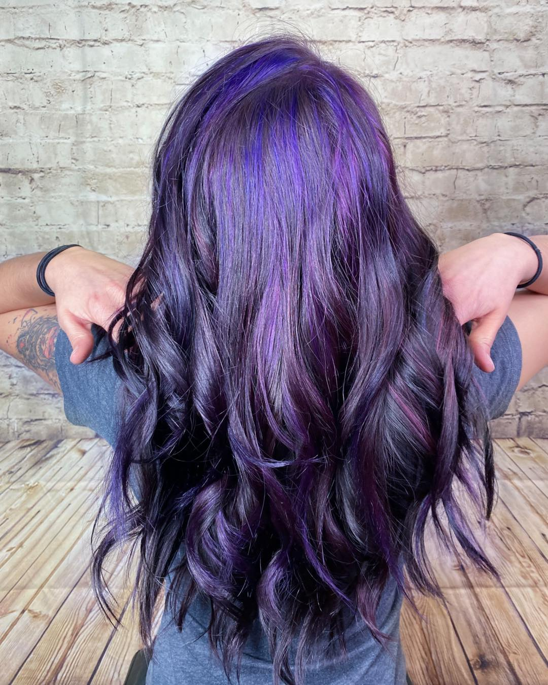 Person with long wavy purple hair viewed from behind against a brick wall.