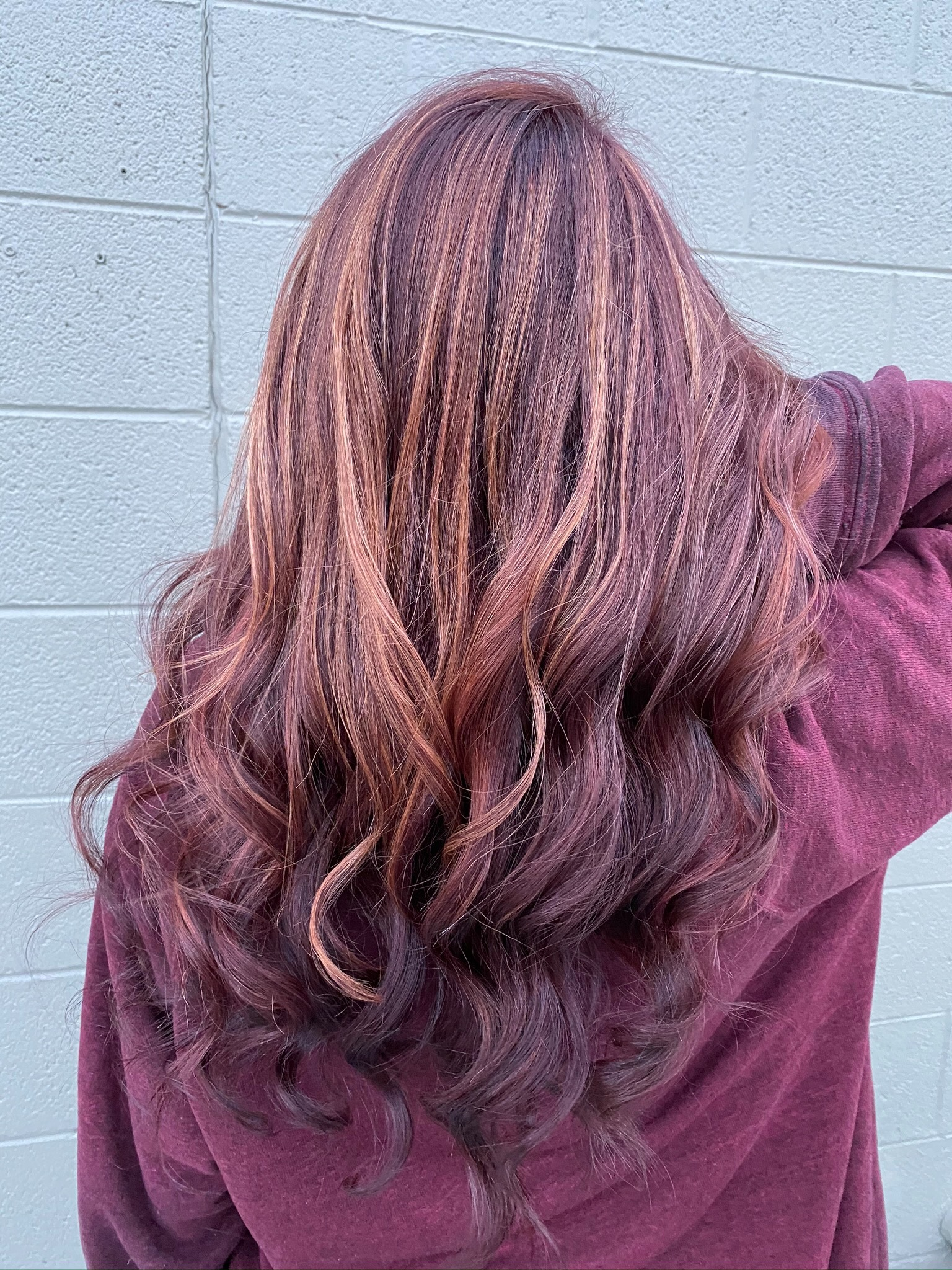 Person with long auburn hair facing a gray tiled wall, wearing a maroon jacket.