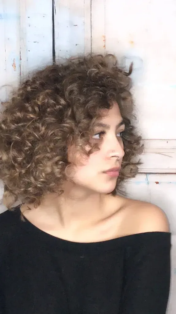 Person with curly hair in a black top looking left against a light wooden wall