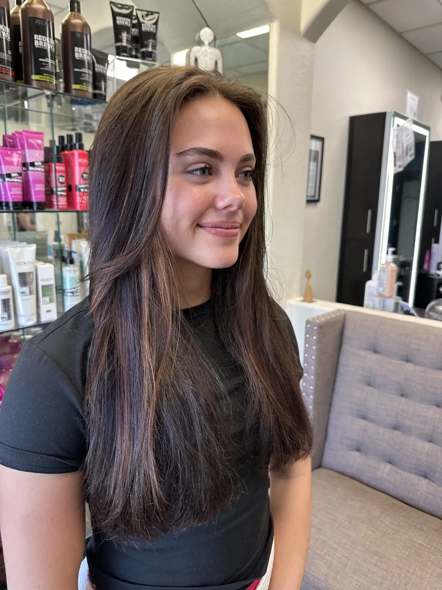 Smiling person with long brown hair seated in a salon, with hair products and styling chairs nearby