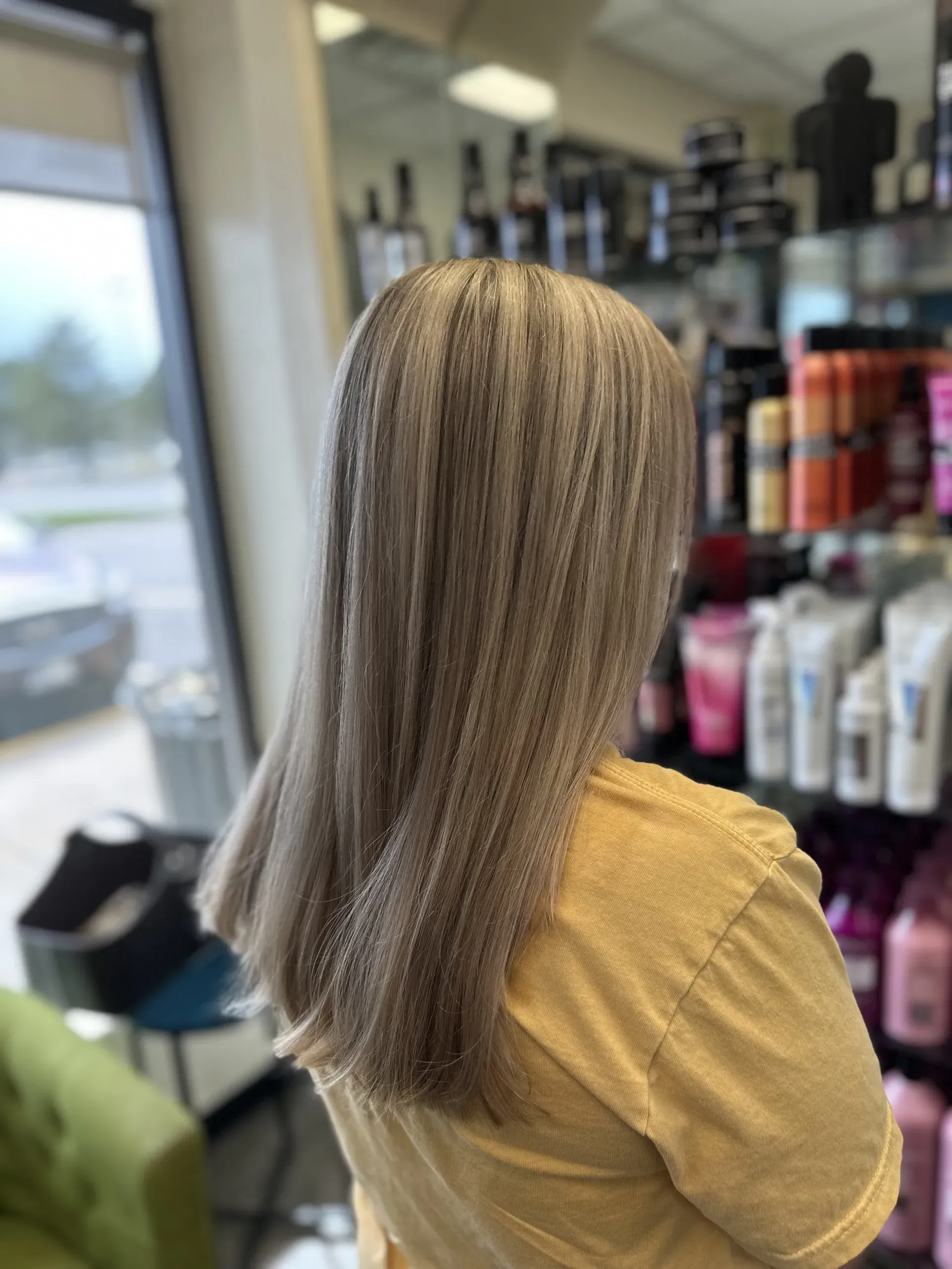 Person with shoulder-length blonde hair in a salon, seen from behind near haircare product shelves.
