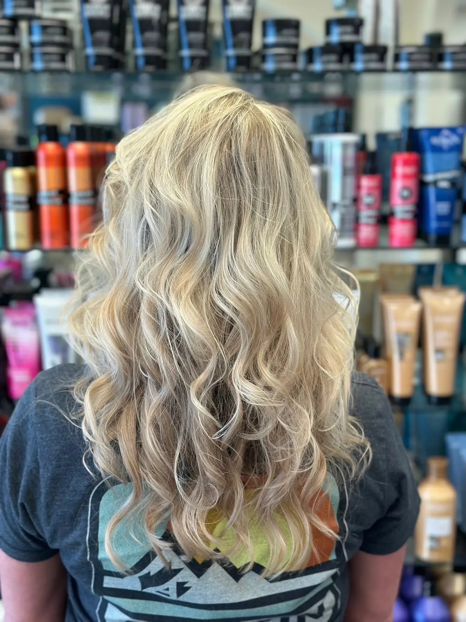 Back view of a person with long wavy blonde hair in front of a wall of hair products