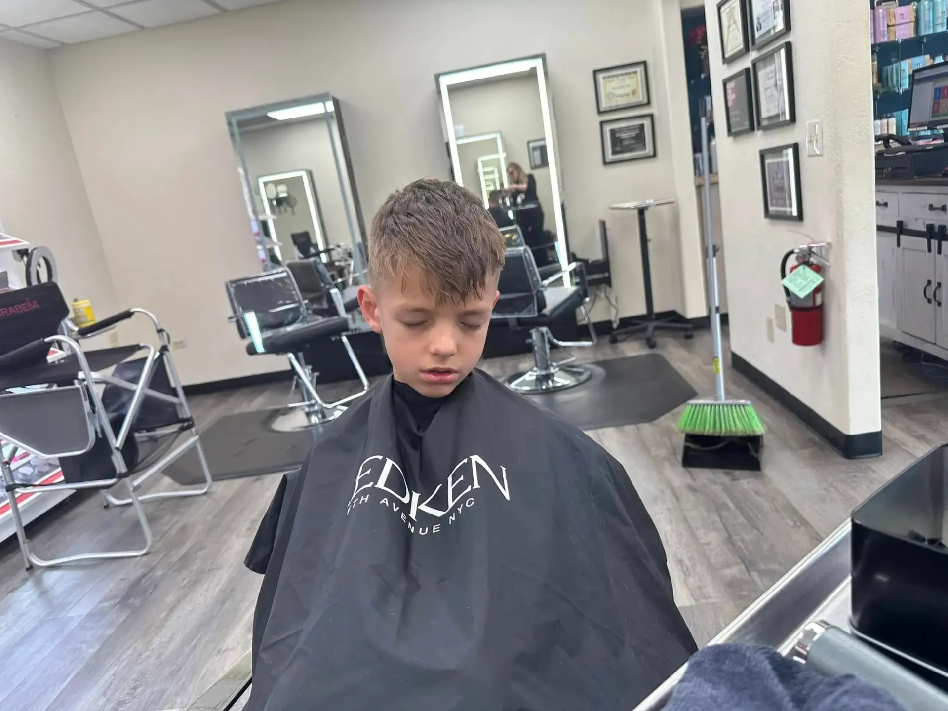 Child in a black salon cape sitting in a barbershop chair, looking down.