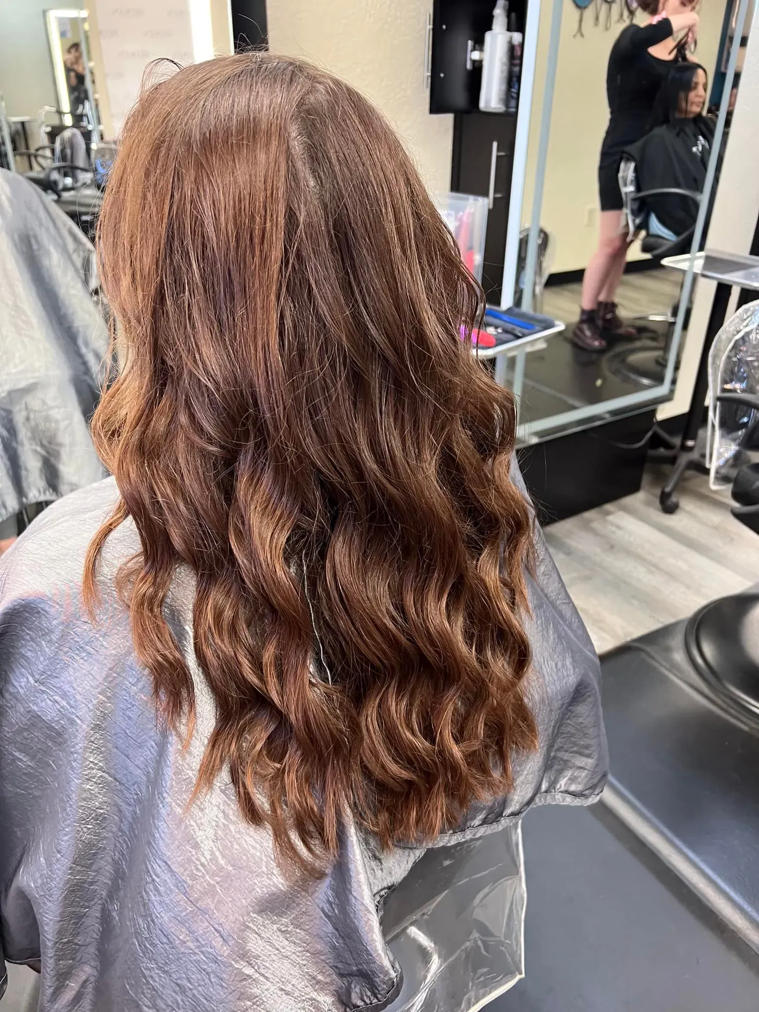 Back view of long, wavy auburn hair in a salon chair, with a mirror and stylist in the background