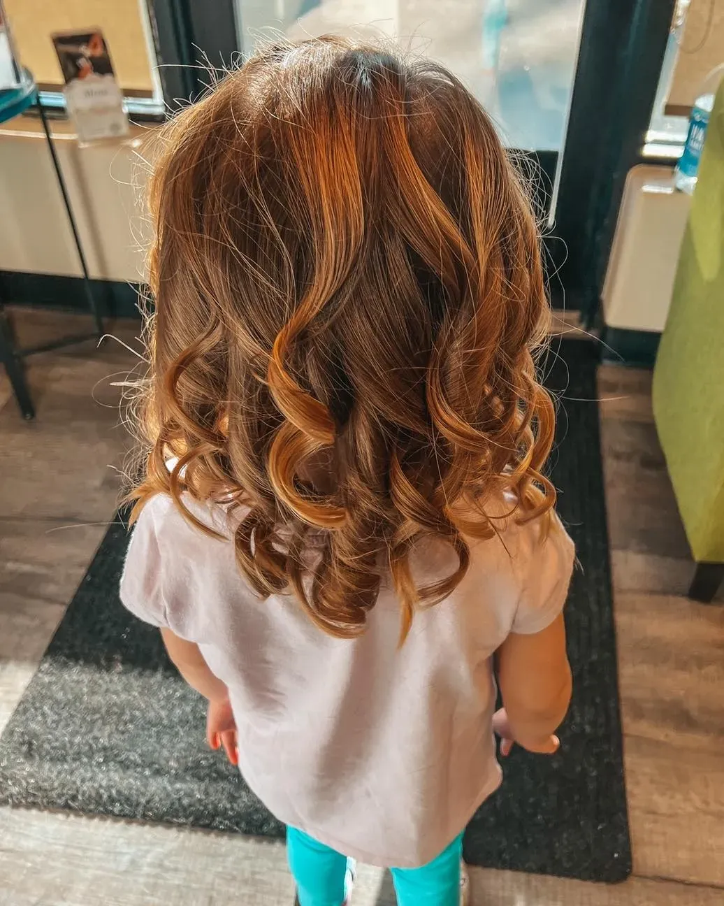 Child with curly brown hair in a salon, seen from behind, wearing a white shirt and teal pants.