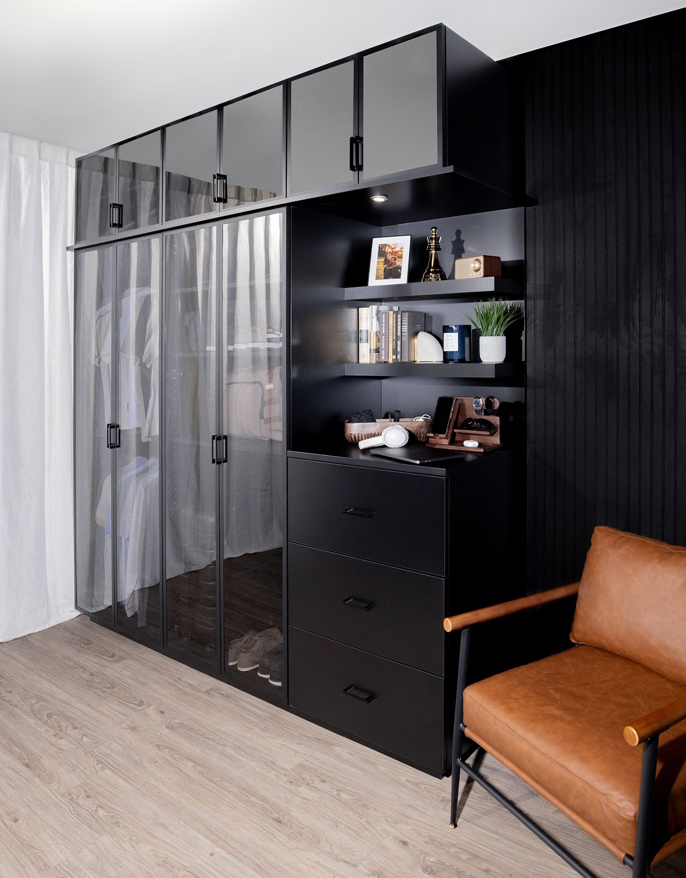 Black wardrobe and shelving unit beside a tan chair in a modern room with wood floor and sheer curtains