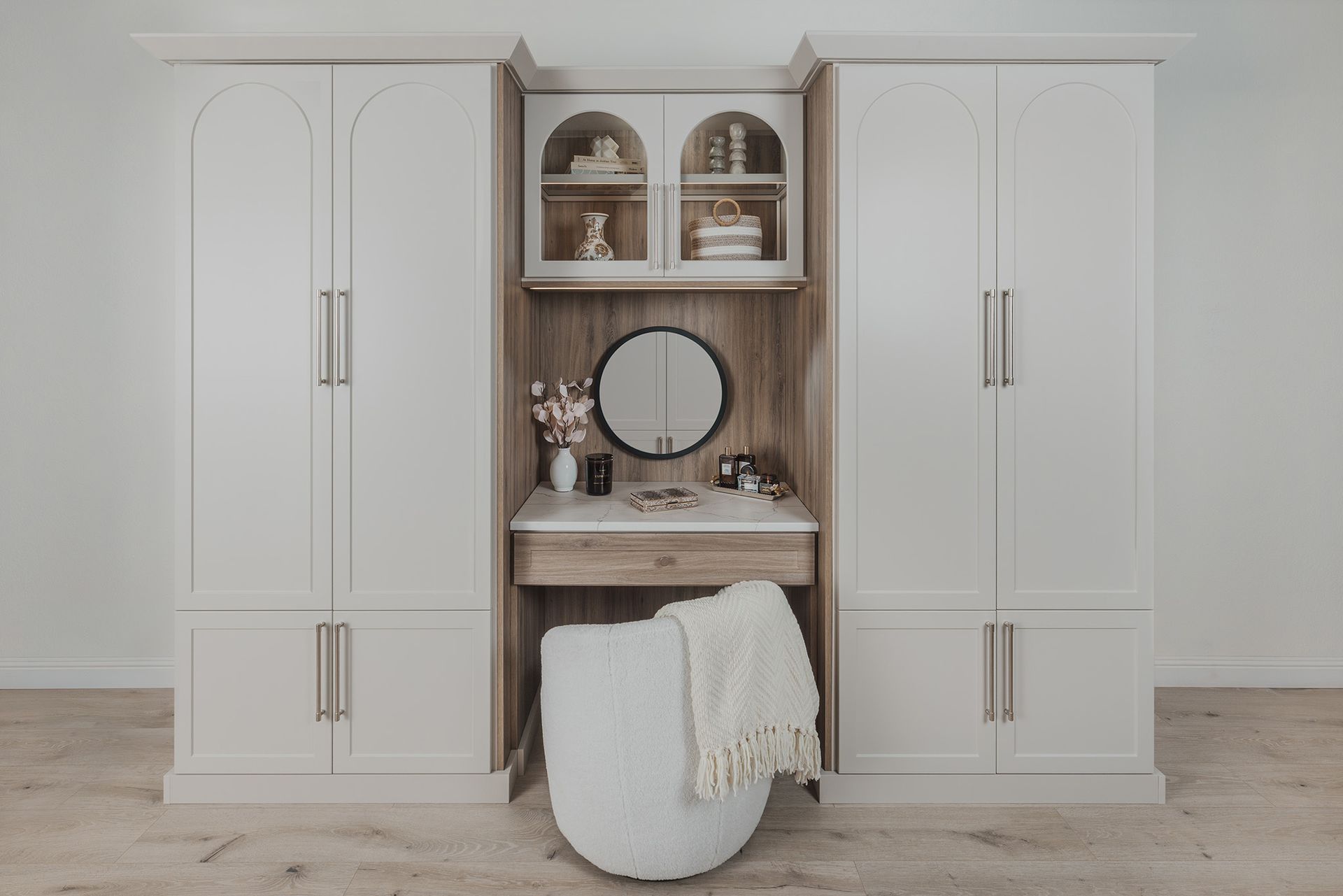 White built-in cabinets framing a vanity with round mirror, decor shelves, and a stool with a cream blanket.