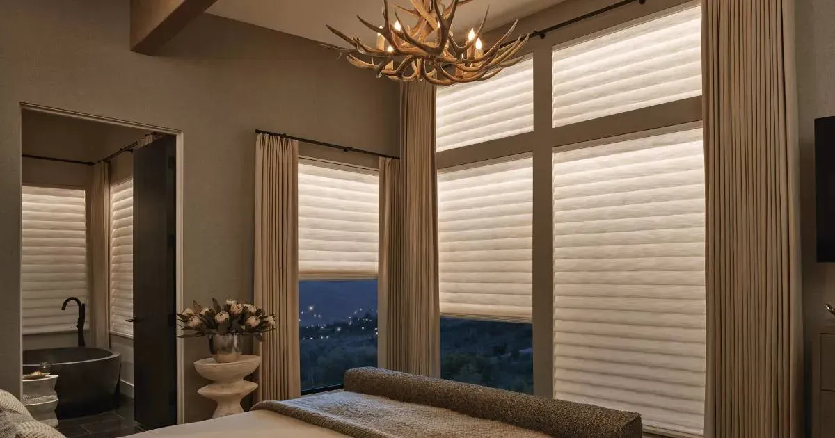A bedroom with a large bed , aura shades, a chandelier , and lots of windows.