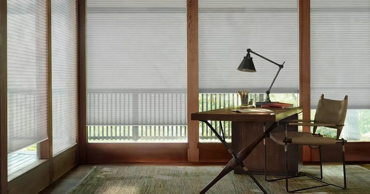 There is cellular shades, a desk and chair in the room with a lamp.