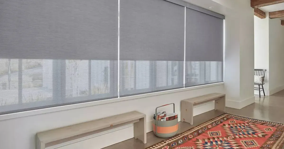 A living room with roller shades a rug , benches , and blinds on the windows.