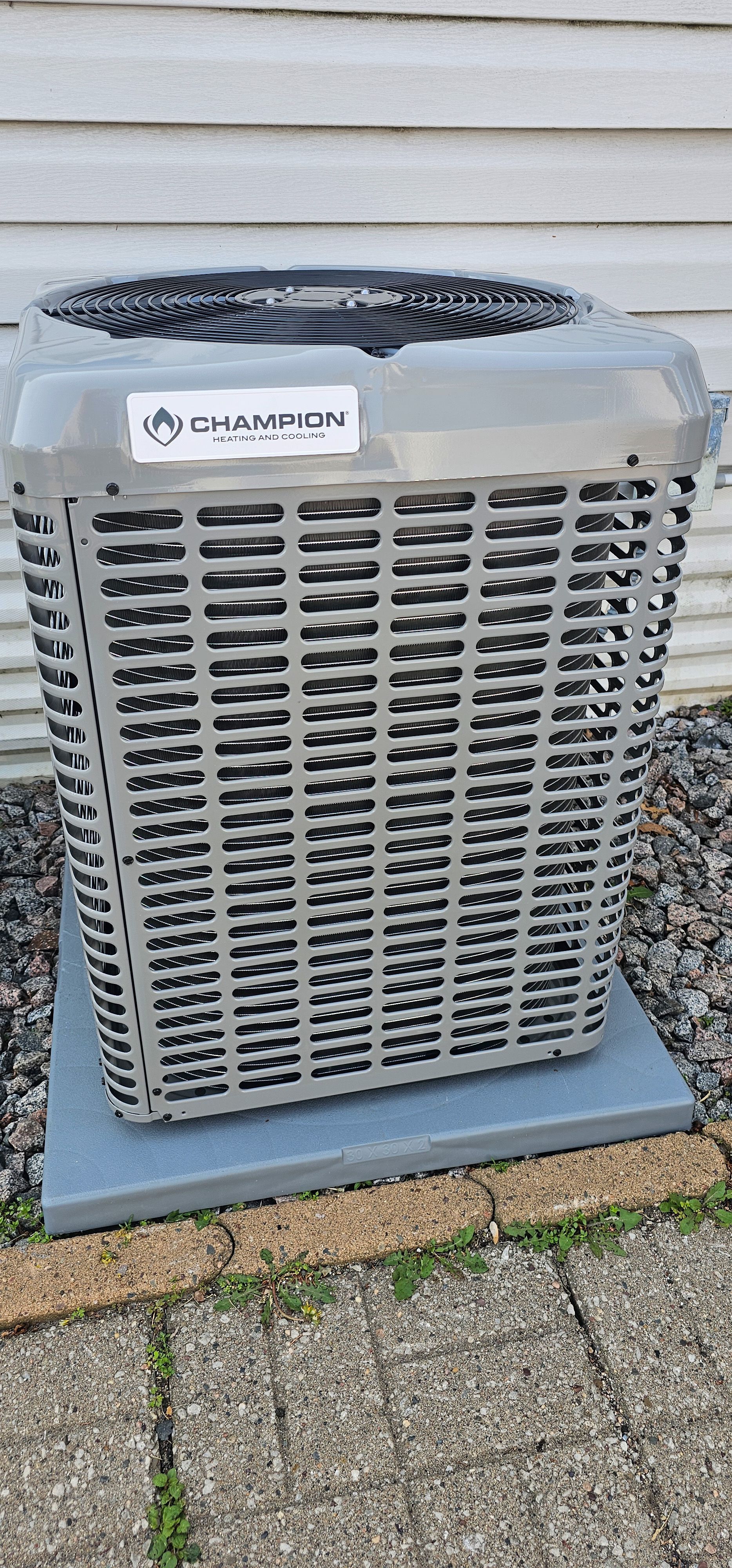 Outdoor air conditioner unit with gray metal grille beside a house wall and patio.