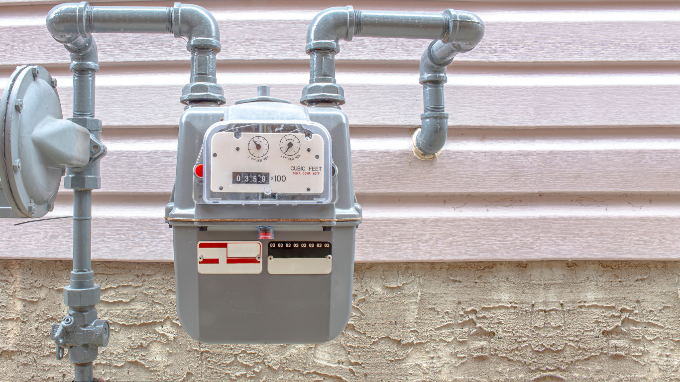 Gas meter with silver pipes mounted on beige house siding and concrete foundation.