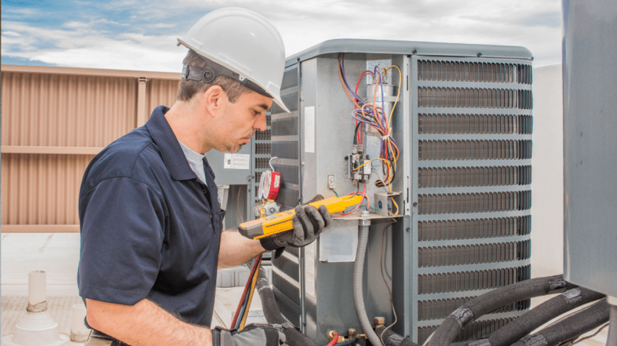 Technician in hard hat using a multimeter to inspect an outdoor HVAC unit.