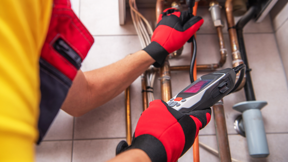 Electrician in red gloves inspecting exposed wiring and pipes with a multimeter in a wall cavity