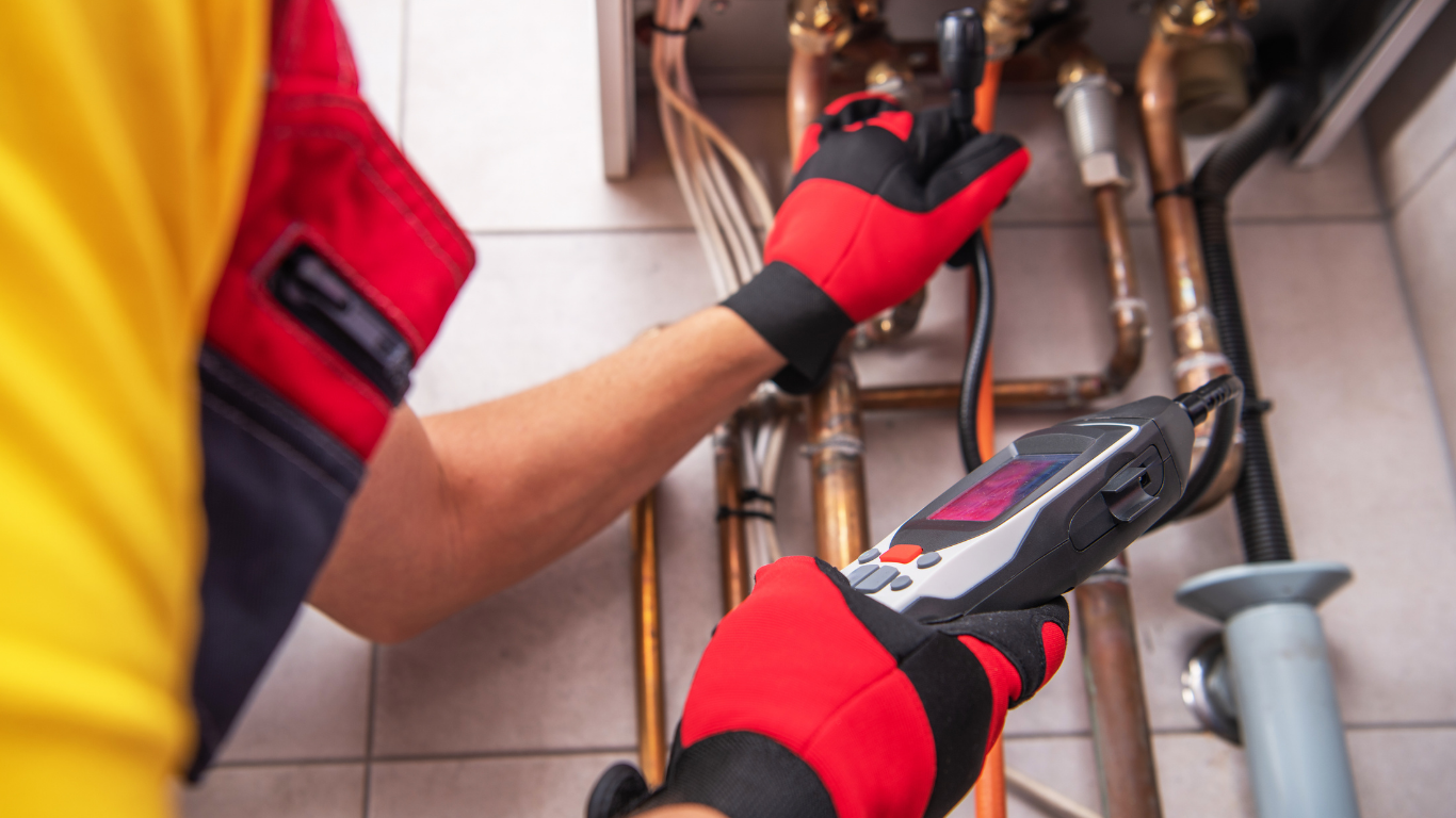 Gloved worker inspecting copper pipes under a sink with a handheld device and wrench